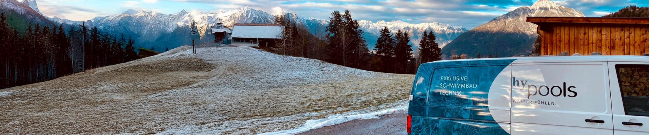 Firmenauto im Vordergrund im Hintergrund schneebedeckte Berge davor ein Haus mit Wald